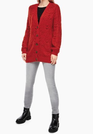 Vrouw die een rood gebreid vest met knopen draagt, lichtgrijze skinny jeans, zwarte enkellaarzen en een zwarte top eronder.