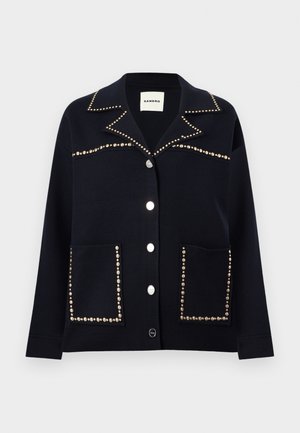 Μαύρο σακάκι με κουμπιά και μυτερό γιακά, διακοσμημένο με ασημένιες καρφίτσες κατά μήκος του γιακά, του στήθους και των άκρων των τσεπών.