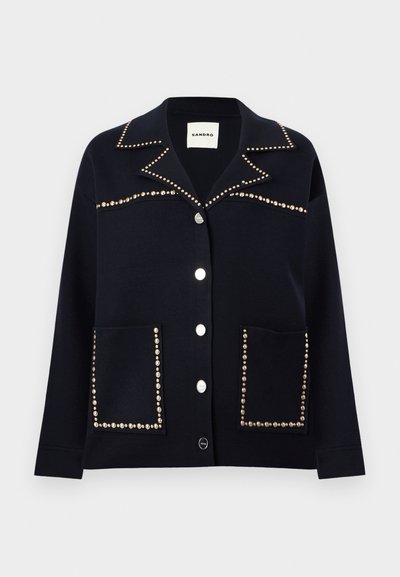 Μαύρο σακάκι με κουμπιά και μυτερό γιακά, διακοσμημένο με ασημένιες καρφίτσες κατά μήκος του γιακά, του στήθους και των άκρων των τσεπών.