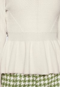 Pull en tricot crème avec texture côtelée, ourlet en peplum et manches longues. Comprend une taille lisse et des détails de conception superposés.