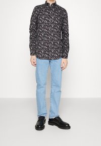 Maglietta nera a maniche lunghe con piccolo motivo floreale, colletto con bottoni, abbinata a jeans azzurri chiari e scarpe nere. Tessuto liscio, taglio aderente.