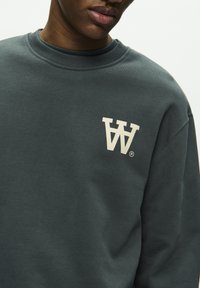 Dunkelgrüner Sweatshirt mit geripptem Kragen, ausgestattet mit einem großen weißen Logo auf der Brust. Hergestellt aus einem weichen, strukturierten Stoff.