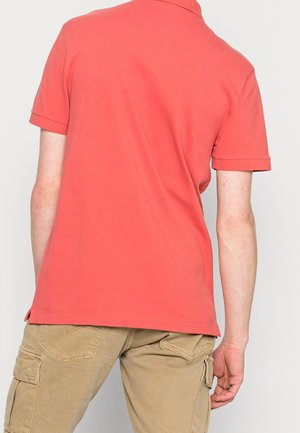 Coral polo shirt with short sleeves, flat collar, and vented hem, paired with light beige cargo pants featuring pockets and textured fabric.