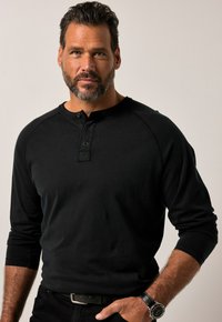 Hombre con cabello corto y oscuro y barba, vistiendo una camiseta Henley de manga larga negra y un reloj de pulsera, de pie contra un fondo claro y liso.