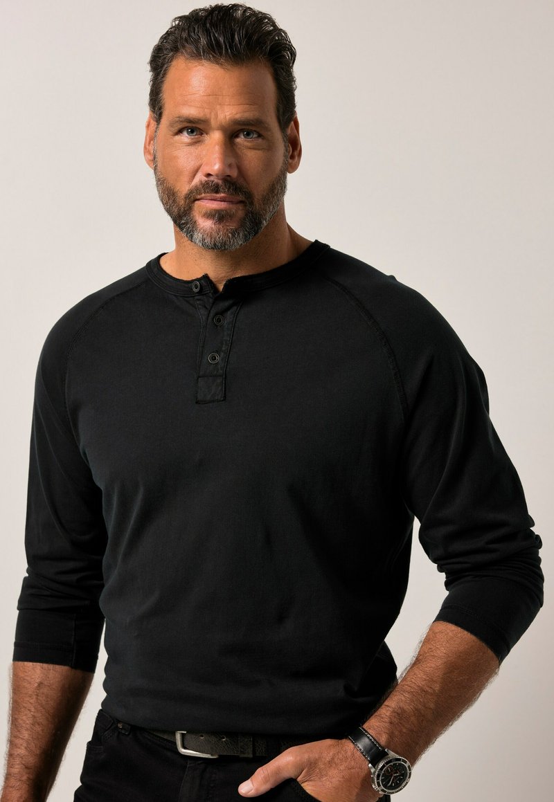 Hombre con cabello corto y oscuro y barba, vistiendo una camiseta Henley de manga larga negra y un reloj de pulsera, de pie contra un fondo claro y liso.