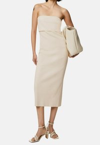 Robe midi beige sans bretelles avec un corsage ajusté, une texture lisse et une silhouette droite. Accordée avec un sac beige texturé et des talons clairs.