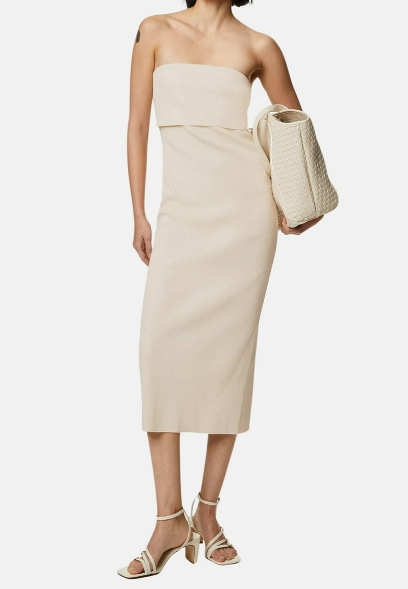 Robe midi beige sans bretelles avec un corsage ajusté, une texture lisse et une silhouette droite. Accordée avec un sac beige texturé et des talons clairs.