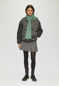 Grau gesteppte Jacke mit schwarzen Fleece-Ärmeln, kombiniert mit einem grünen Schal, einem gestreiften grauen Rock, schwarzen Strumpfhosen und massiven schwarzen Stiefeln.