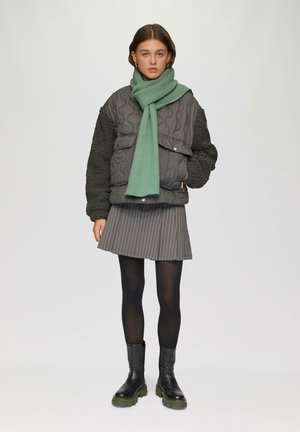 Gequilted grijze jacket met zwarte fleece mouwen, gecombineerd met een groene sjaal, een gestreepte grijze rok, zwarte panty’s en stevige zwarte laarzen.