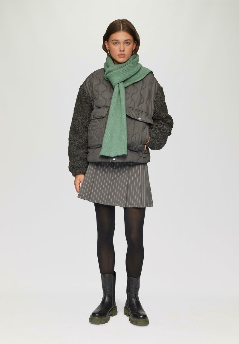 Gequilted grijze jacket met zwarte fleece mouwen, gecombineerd met een groene sjaal, een gestreepte grijze rok, zwarte panty’s en stevige zwarte laarzen.