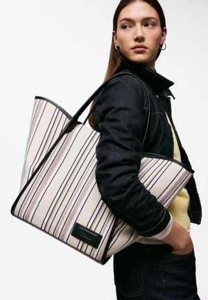 Giovane donna con giacca di jeans scura che porta una grande borsa beige con strisce verticali nere e rosa sulla spalla.