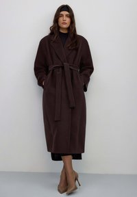 Lange, donkerbruine wollen jas met ceintuur, brede kraag en lange mouwen. Model staat met handen in de zakken, draagt beige hakken. Minimalistisch ontwerp.