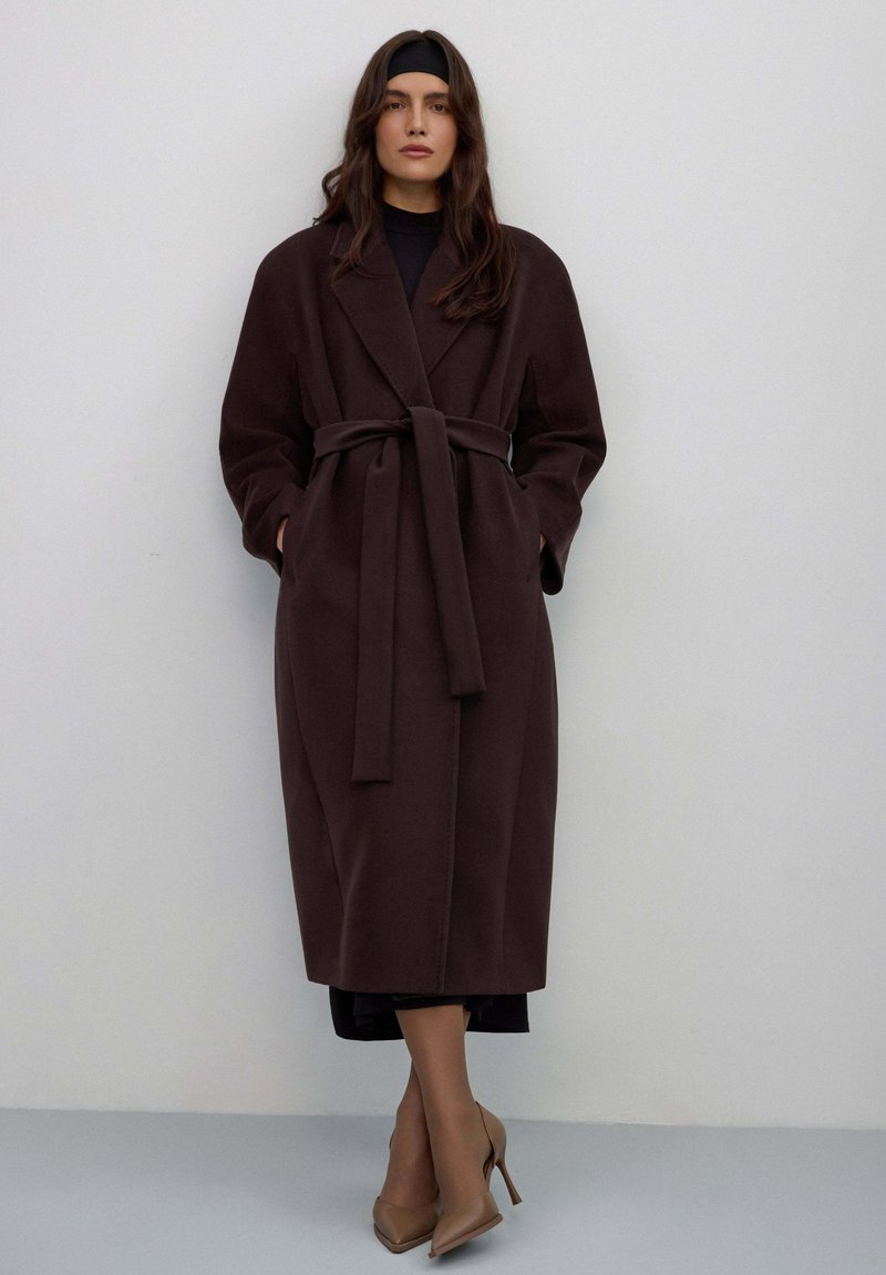 Lange, donkerbruine wollen jas met ceintuur, brede kraag en lange mouwen. Model staat met handen in de zakken, draagt beige hakken. Minimalistisch ontwerp.