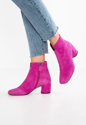 Persoon die felroze suède enkellaarsjes met blokhakken draagt en lichtblauwe cropped jeans met rafelige zoom, tegen een effen witte achtergrond.