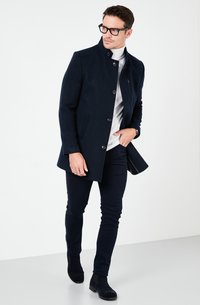 Cappotto in lana blu navy con colletto alto, chiusura a bottoni e tasche laterali. Indossato sopra un dolcevita bianco, abbinato a pantaloni scuri e stivaletti neri.