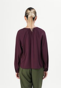 Woman with blonde ponytail wearing a long-sleeve dark purple blouse and olive green pants, shown from behind against a plain background.