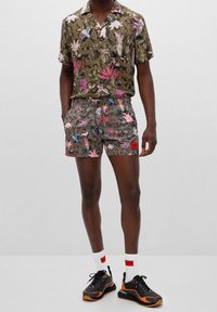 Mann in passendem olivgrünem, blumengem Kurzarmhemd und Shorts, mit schwarz-orangenen Sneakers und weißen Socken mit rotem Logo.