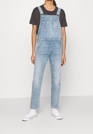 Denim overalls in light blue with an adjustable strap, front pocket, and gold-tone hardware. Worn over a black t-shirt, paired with white sneakers.