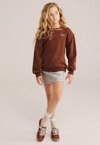 Rjav sweatshirt s izvezenim besedilom, siva kavbojka, bež in bela prugasta nogavica ter bordo superge z vezalkami.