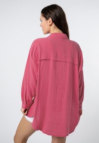 Lange mouwen, knoopsluitende blouse van gemêleerd felroze stof, met een rugpas en een ontspannen pasvorm, zichtbaar vanaf de achterkant.