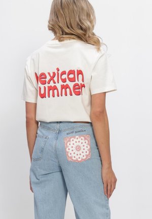 Blond persoon die een gebroken witte t-shirt draagt met rode tekst "Mexican Summer" en lichtblauwe spijkerbroek met een patroonlapje op de achterzak.