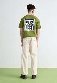 T-shirt vert olive à manches courtes avec un graphique noir "OBEY" au dos, associé à un pantalon cargo crème et des bottes noires. Texture de tissu lisse.