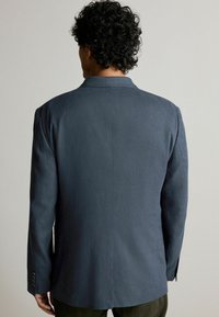 Dunkelblazer mit strukturiertem Design, seitlichem Rückenschlitz und geknöpften Ärmeln. Der Stoff wirkt strukturiert mit einer glatten Oberfläche.