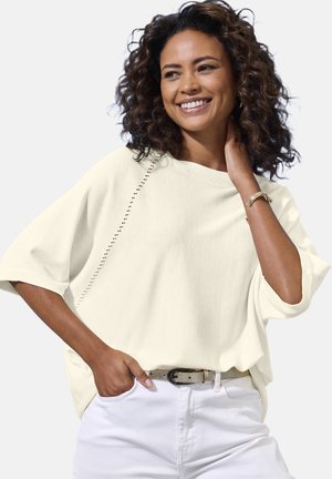 Glimlachende vrouw met krullend haar, gekleed in een crème kleurige gebreide top en witte broek met een lichte riem, hand in de zak, tegen een effen achtergrond.