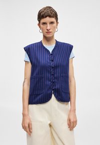 Tamsiai mėlyna liemenė su vertikaliomis rožinėmis juostelėmis, be rankovių, dviem priekinėmis kišenėmis ir penkiais mygtukais. Dėvima virš šviesiai mėlynos marškinių.