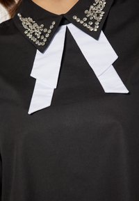 Sort bluse med hvid krave, der har to båndsløjfer, prydet med funklende rhinsten på kraven, glat tekstur og tætsiddende design.