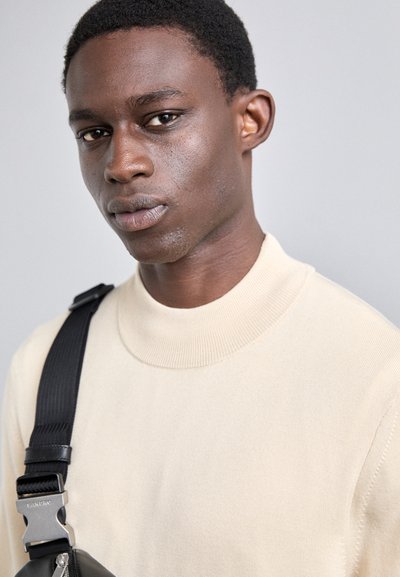 Heller beige Pullover mit hohem Kragen und gerippter Textur, kombiniert mit einer schwarzen Schultertasche mit silbernen Beschlägen. Hintergrund ist neutral grau.