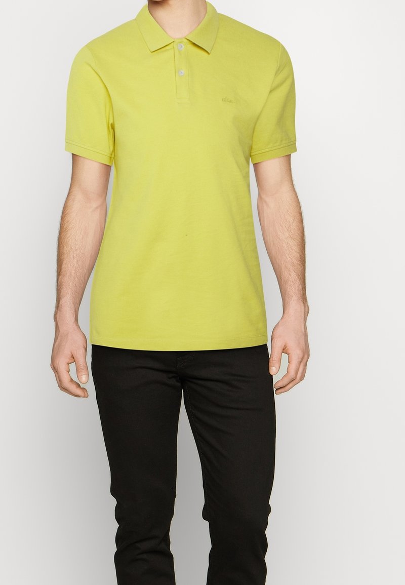 Polo jaune vif en coton, doté d'un col classique, de manches courtes et d'un logo discret sur la poitrine. Porté avec un pantalon noir.