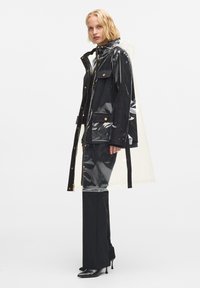 Chaqueta de lluvia oversize negra y transparente con herrajes dorados, múltiples bolsillos y capucha ajustable. Textura suave y brillante.