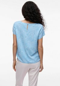 Ljusblå blus med ett prickigt vitt mönster, korta ärmar, rundad nederkant och en nyckelhålsglipa på baksidan. Tillsammans med ljusa byxor.