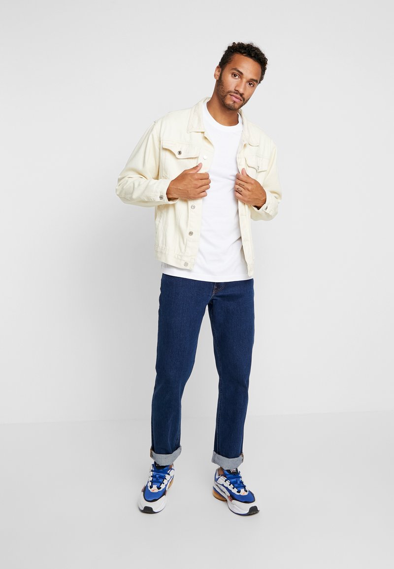 Lichtbeige denimjas, witte t-shirt, donkerblauwe opgerolde jeans en blauwe sneakers met witte en bruine accenten, poserend tegen een effen achtergrond.