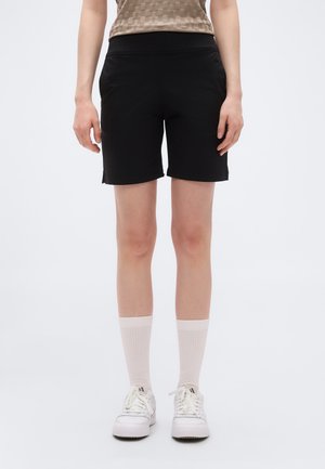 Osoba oblečená v černých šortkách do kolen, béžových ponožkách, bílých teniskách a béžovém vzorovaném topu bez rukávů, stojící na bílém pozadí.