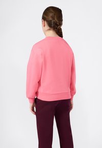 Mulher com cabelo castanho trançado, vestindo um suéter rosa brilhante e calças burgundy escuras, de pé de costas contra um fundo branco simples.