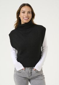 Čierny pletený sveter s vysokým golierom a rebrovaným lemom, nosený cez bielu košeľu s dlhým rukávom, skombinovaný so svetlosivými džínsami.