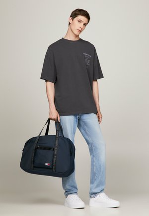 Borsa a tracolla blu navy con cinture nere caratterizzata da un logo. Indossata con una t-shirt grigia oversize e jeans azzurri chiari. Sneakers bianche.