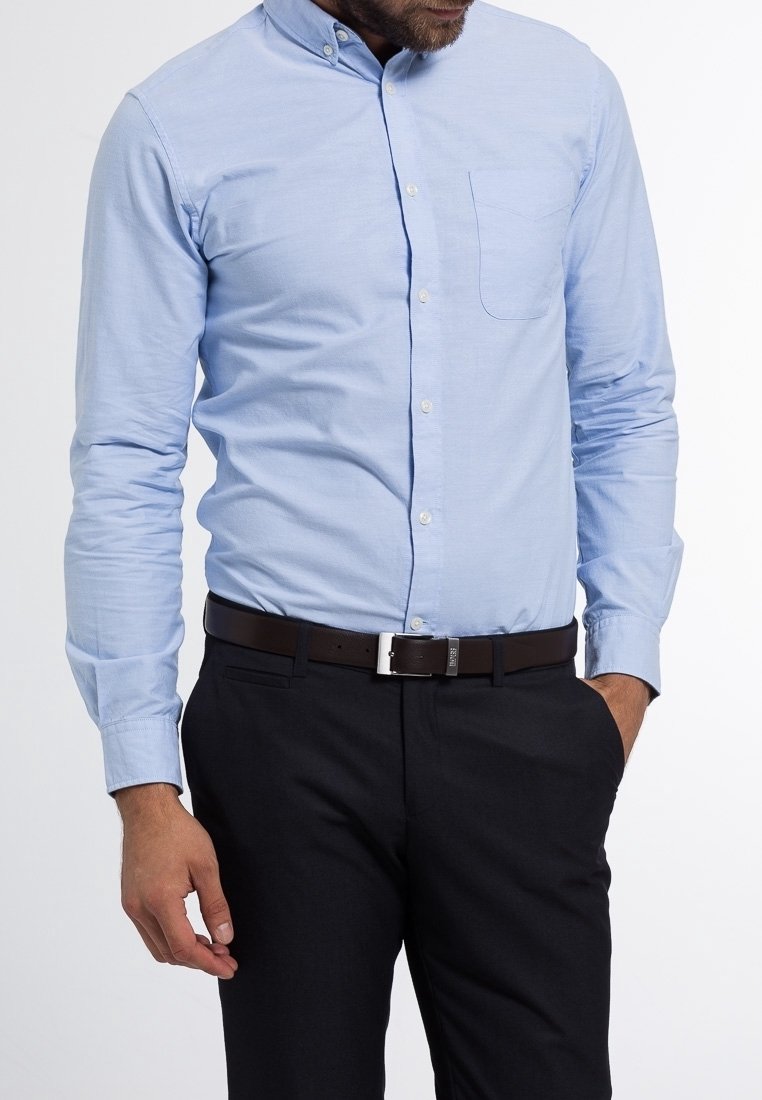 Homme portant une chemise bleu clair boutonnée rentrée dans un pantalon noir avec une ceinture noire, debout avec une main dans la poche.