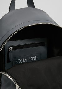 Sac à dos en cuir gris avec fermeture éclair métallique, présentant un intérieur noir et une poche de marque avec zip. Texture lisse et forme arrondie.