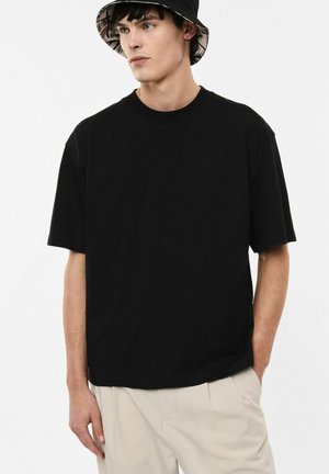 Jonge man met een zwarte bucket hat, oversized zwart T-shirt en beige broek, staand met één hand in zijn zak, kijkt opzij.