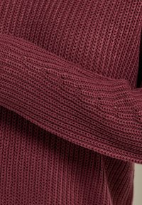 Gestrickter Pullover in tiefem Burgunderrot mit einem strukturierten, gerippten Muster. Die Ärmel sind lang und leicht übergroß und sorgen für ein gemütliches Gefühl.