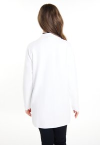 Lungo cardigan bianco con una texture liscia e una vestibilità rilassata, caratterizzato da maniche lunghe e orlo arrotondato. Lo sfondo è bianco uniforme.