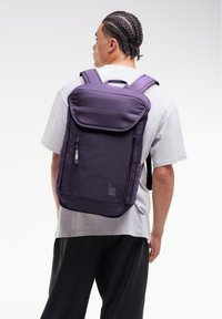 Sac à dos en tissu violet avec des bretelles rembourrées, de forme rectangulaire, fermeture à rabat à l'avant, poche latérale et détails de logo.