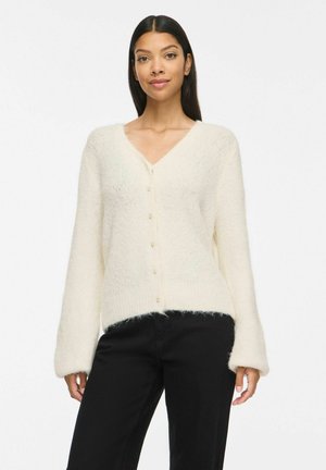 Femme aux longs cheveux foncés portant un cardigan blanc doux boutonné et un pantalon noir sur un fond clair uni.