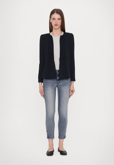 Marineblauwe jas met geplooide voorkant, grijze top eronder en lichtblauwe skinny jeans. Model draagt zwarte ballerina's; minimalistisch, gestructureerd ontwerp.