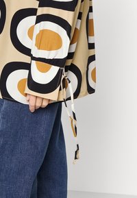 Camisa de mangas longas em bege com grandes padrões circulares em preto e laranja. Apresenta um cordão nos punhos e é combinada com jeans azul.