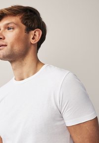 Giovane uomo con pelle chiara e occhi azzurri che indossa una semplice t-shirt bianca a maniche corte, guardando leggermente a sinistra su uno sfondo beige uniforme.