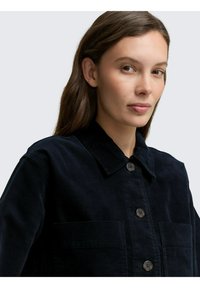 Navy corduroy jas met een klassieke kraag, twee voorzakken en donkere knopen. De textuur van de stof voegt diepte toe aan het ontwerp.